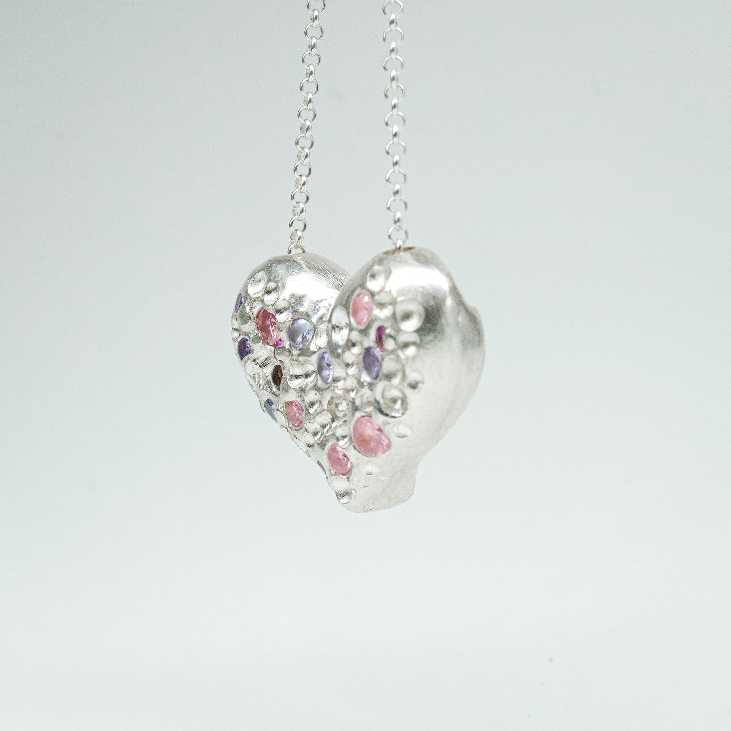 Bulbous heart-shaped sterling silver pendant, hand-sculpted with drilled textures and encrusted with small scattered gemstones. Playful and raw, with a cartoon-like silhouette.