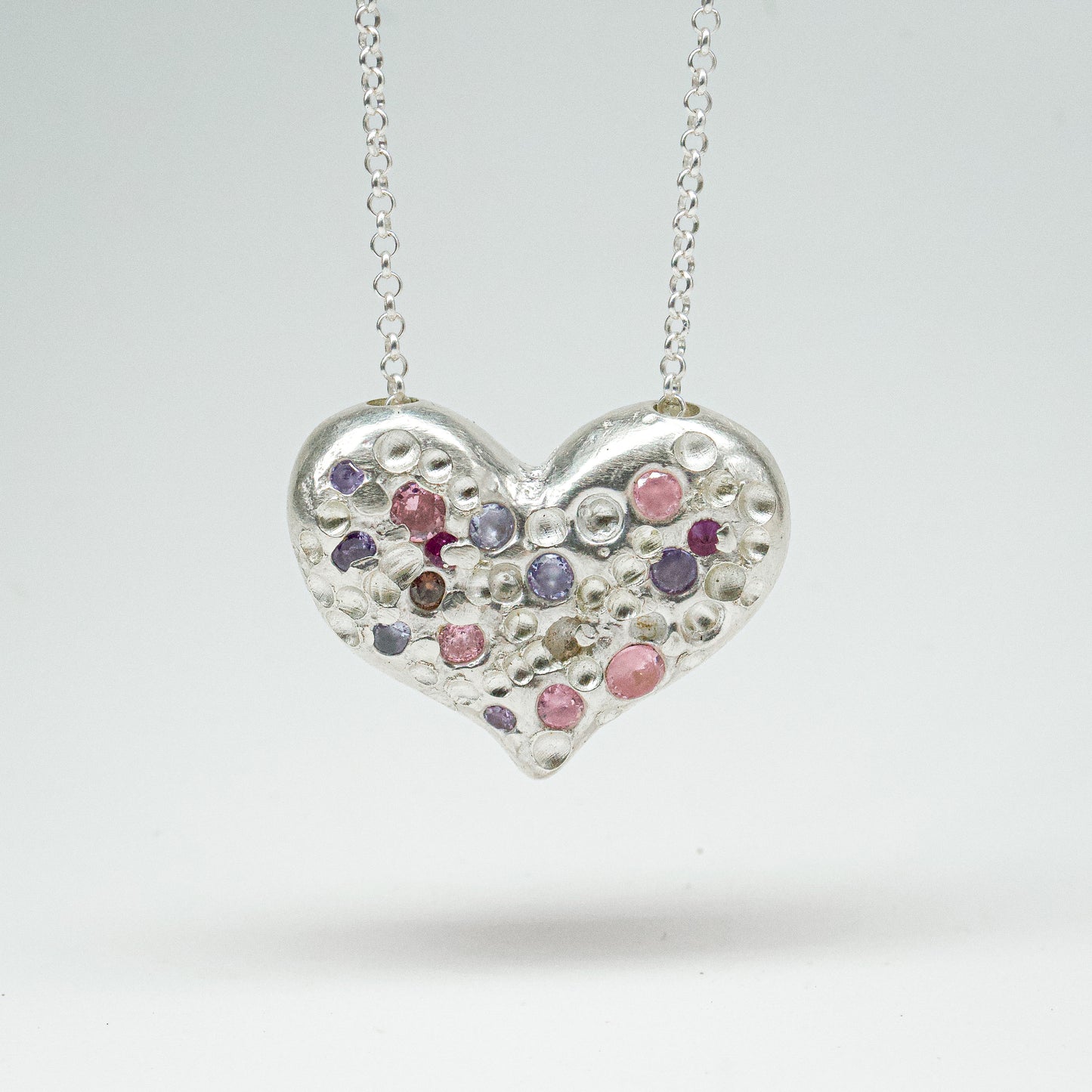 Bulbous heart-shaped sterling silver pendant, hand-sculpted with drilled textures and encrusted with small scattered gemstones. Playful and raw, with a cartoon-like silhouette.