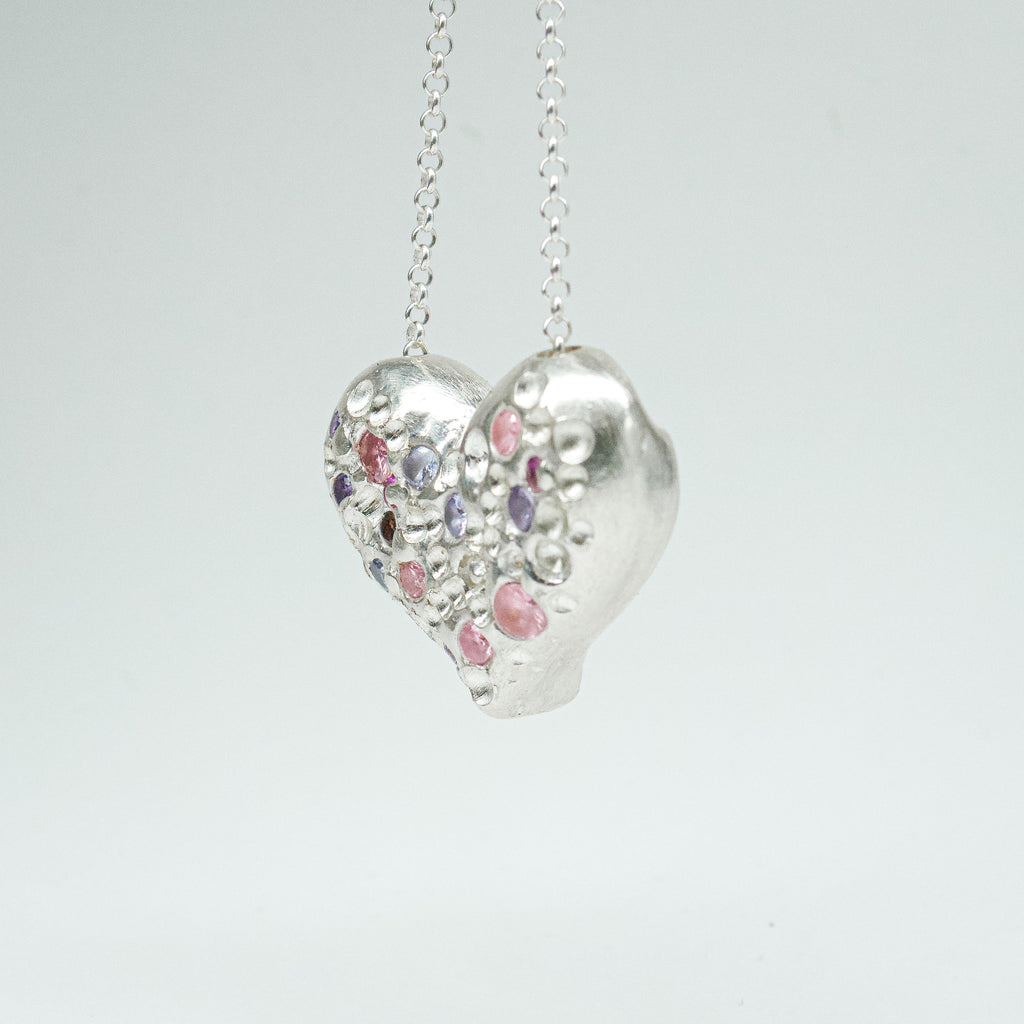 Bulbous heart-shaped sterling silver pendant, hand-sculpted with drilled textures and encrusted with small scattered gemstones. Playful and raw, with a cartoon-like silhouette.