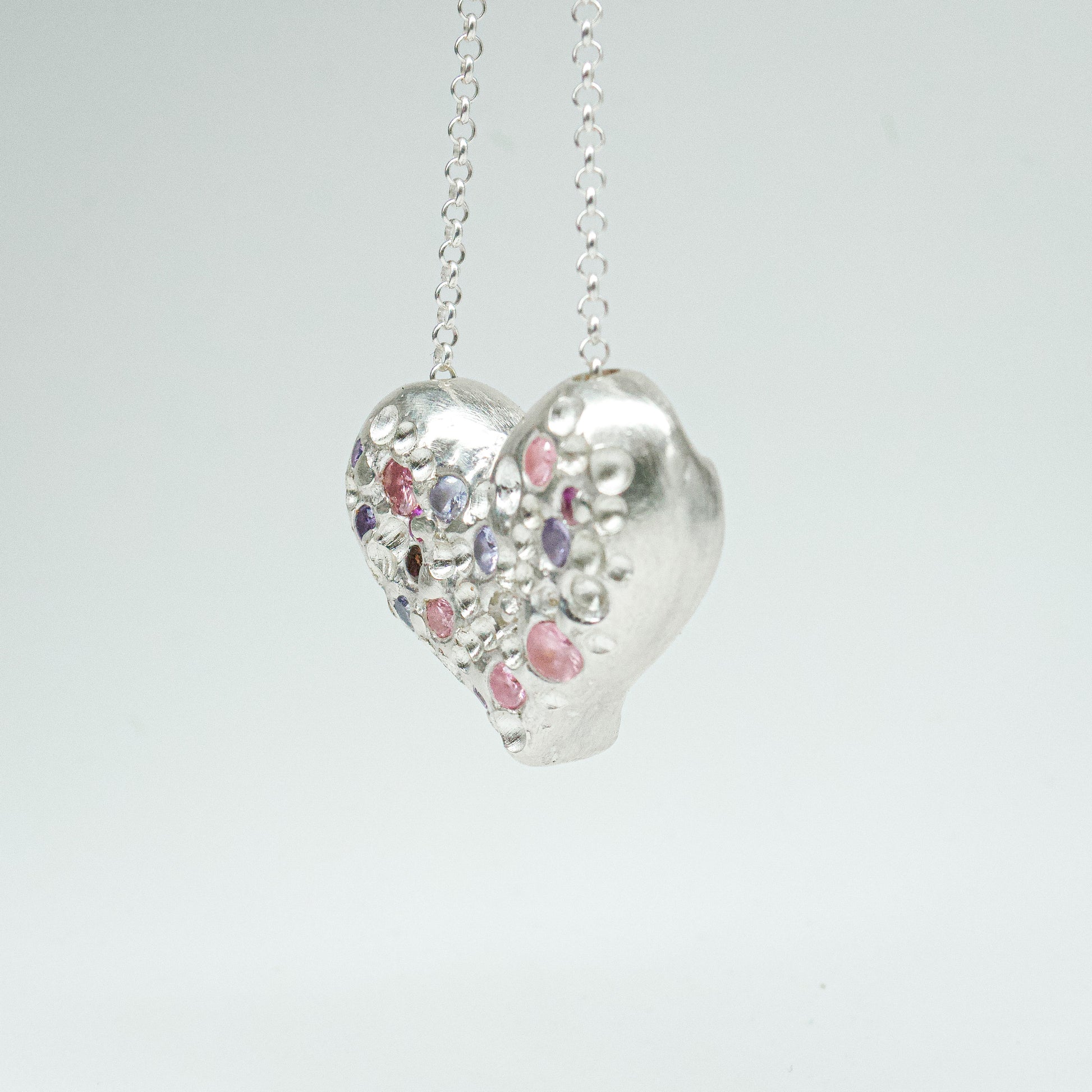 Bulbous heart-shaped sterling silver pendant, hand-sculpted with drilled textures and encrusted with small scattered gemstones. Playful and raw, with a cartoon-like silhouette.