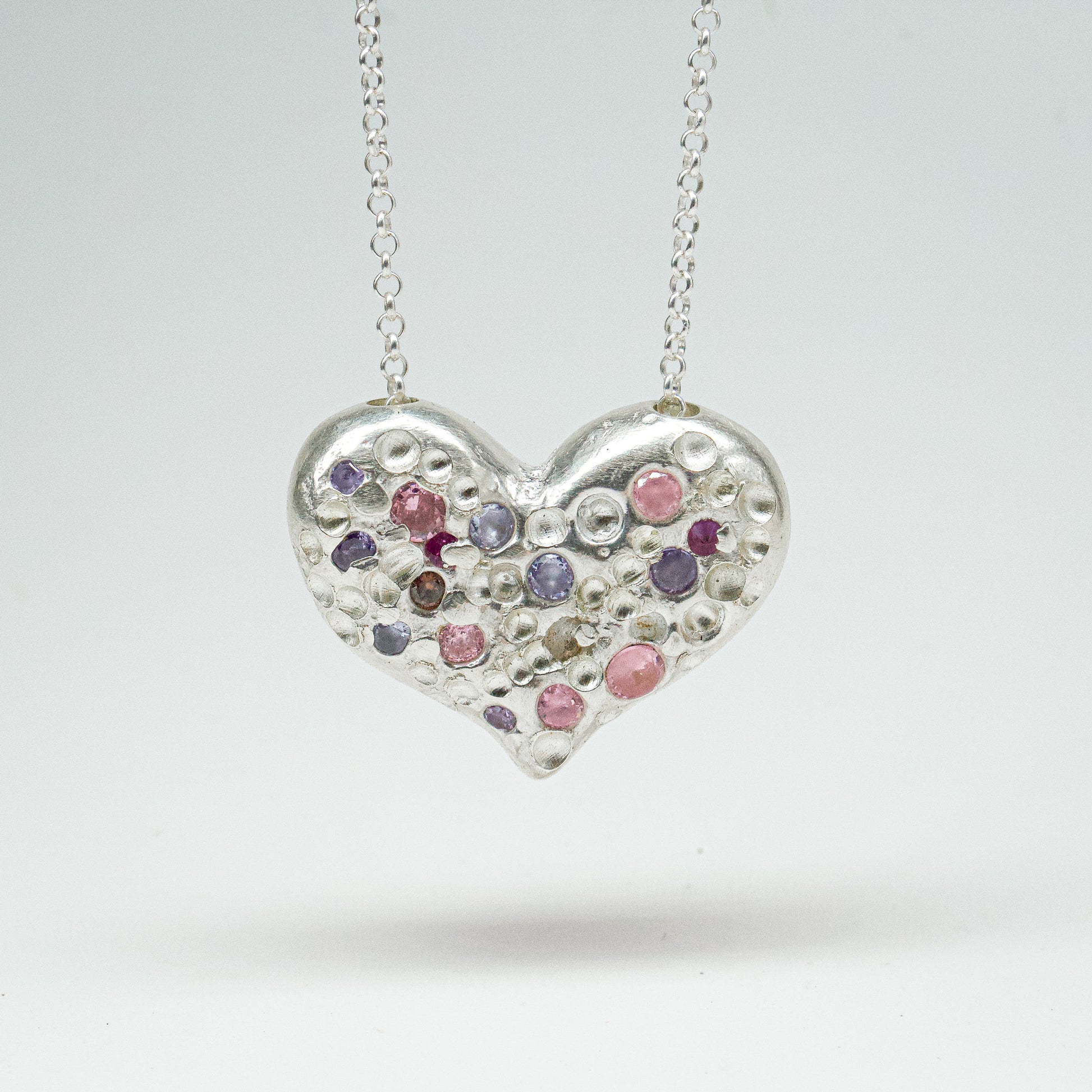 Bulbous heart-shaped sterling silver pendant, hand-sculpted with drilled textures and encrusted with small scattered gemstones. Playful and raw, with a cartoon-like silhouette.