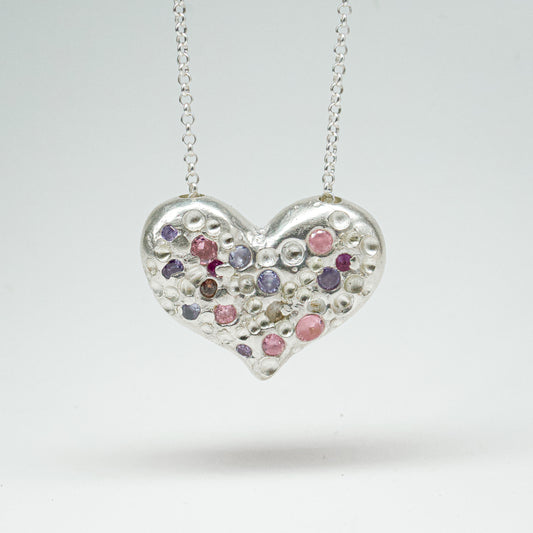 Bulbous heart-shaped sterling silver pendant, hand-sculpted with drilled textures and encrusted with small scattered gemstones. Playful and raw, with a cartoon-like silhouette.